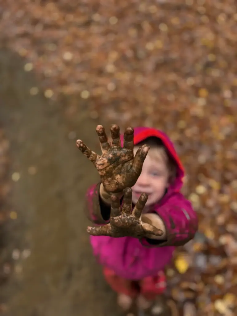 Child holding up mud-covered hands during nature-based education in Vermont at Field, Farm & Forest